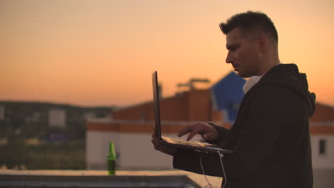 A-man-in-large-white-headphones-types-with-his-fingers-on-the-keyboard-of-a-laptop-standing-on-the-roof-of-a-building-at-sunset-against-the-background-of-the-city.